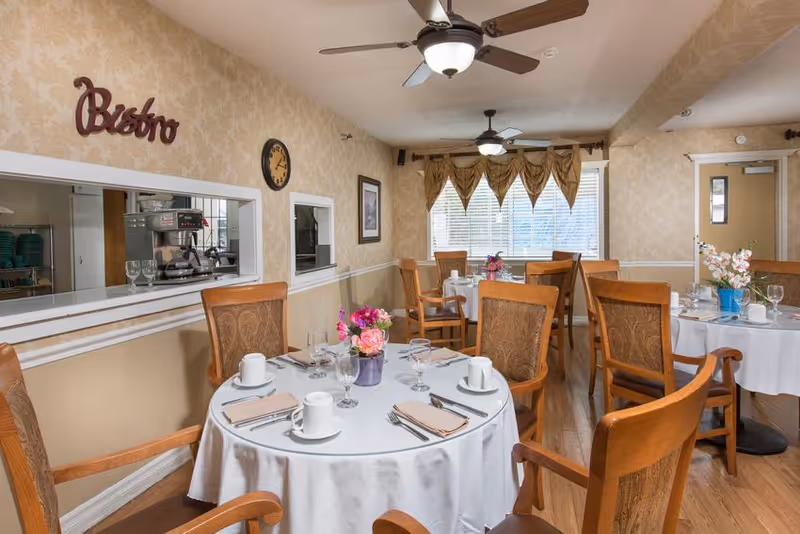 A cozy dining room in a senior living facility with round tables covered in white tablecloths, set with cups, glasses, napkins, and silverware. Each table has a small floral centerpiece. Wooden chairs with cushioned seats surround the tables. The room has beige patterned wallpaper, ceiling fans with lights, and a window with decorative curtains. A serving window labeled 'Bistro' opens to the kitchen area.