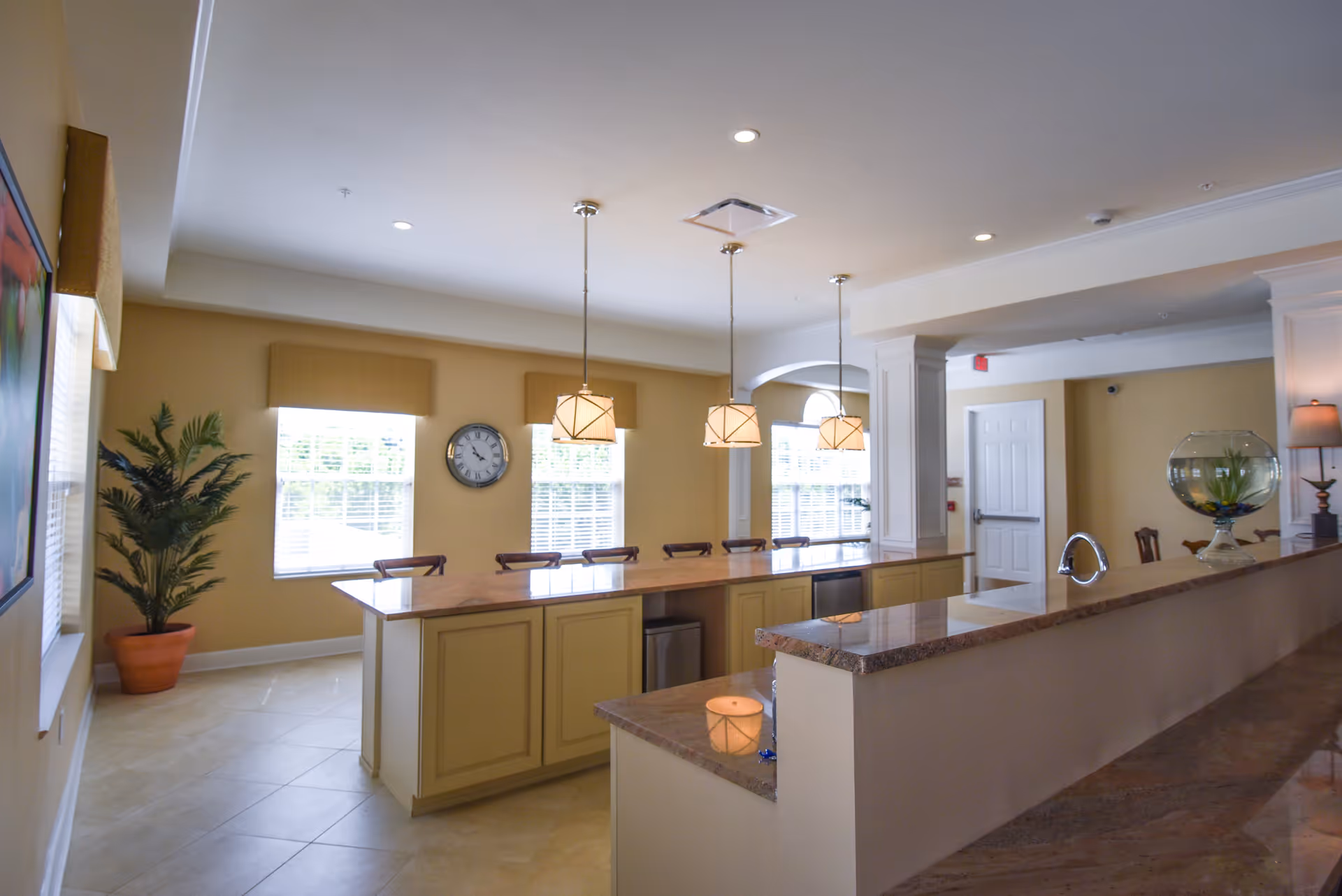 Bright and spacious kitchen area with a long island countertop featuring several bar stools. Three pendant lights hang above the island. There are large windows with blinds and valances letting in natural light. A clock is mounted on the wall between the windows. A potted plant is placed in the corner near the windows. The kitchen has beige walls and tiled floors, with granite countertops and cabinetry.