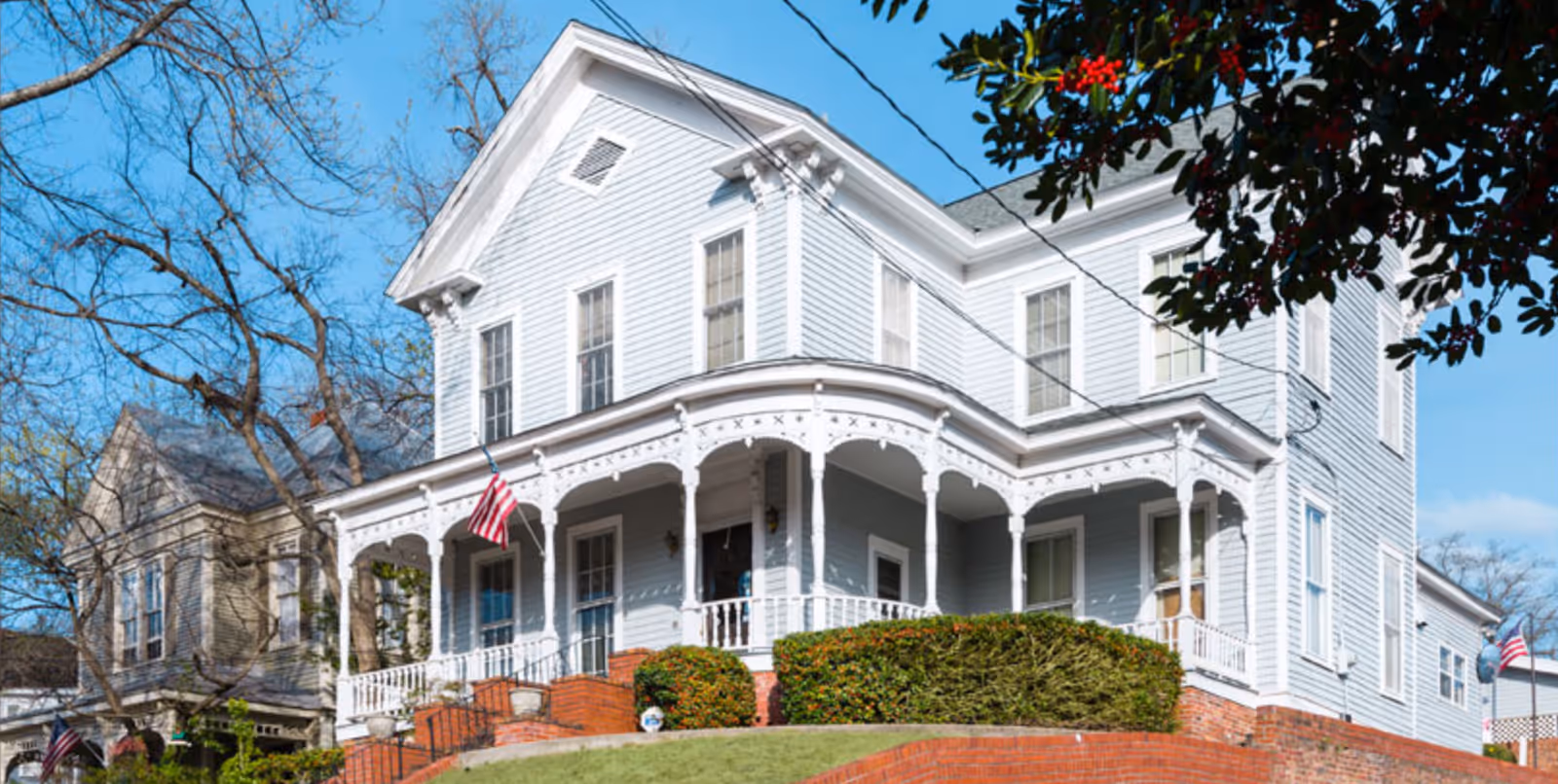 Two-story white Victorian-style building with a wraparound porch, American flag, and brick steps on a grassy hill.