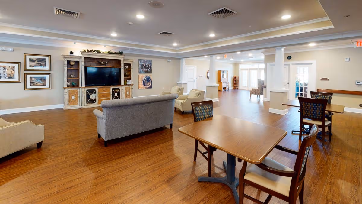 A spacious senior living facility common area with wooden flooring, several tables and chairs, a gray couch, beige armchairs, and a large entertainment center with a flat-screen TV. The walls are decorated with framed pictures and artwork, and the room is well-lit with recessed ceiling lights.