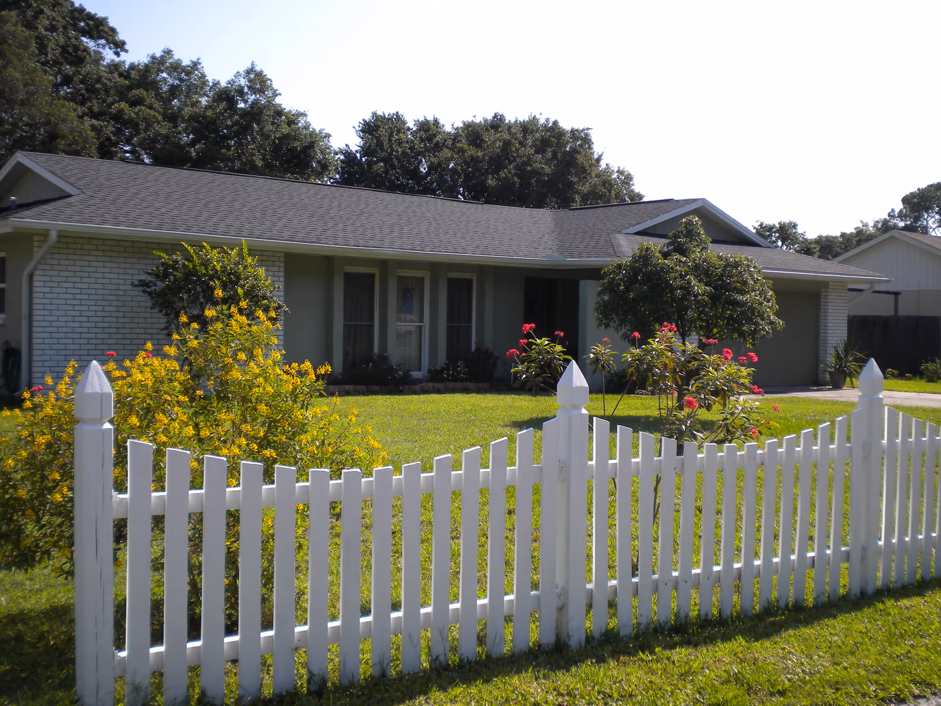 Single-story house with a gray roof and light-colored brick exterior, surrounded by a white picket fence. The front yard has green grass, yellow flowering bushes, and a small tree with pink flowers. Trees are visible in the background under a clear sky.