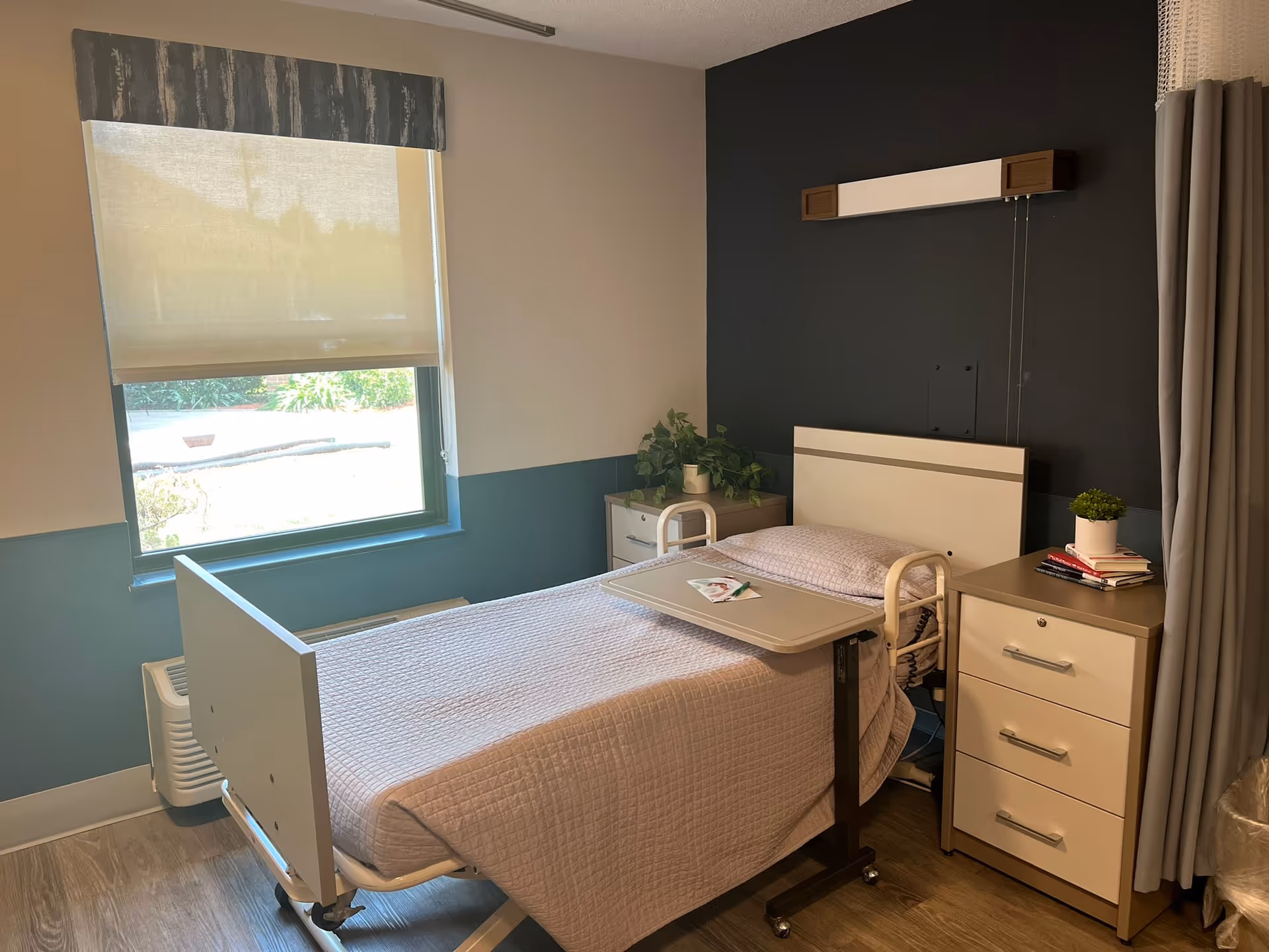 Patient bedroom in a rehab center with a bed, nightstands, plants, and a window.