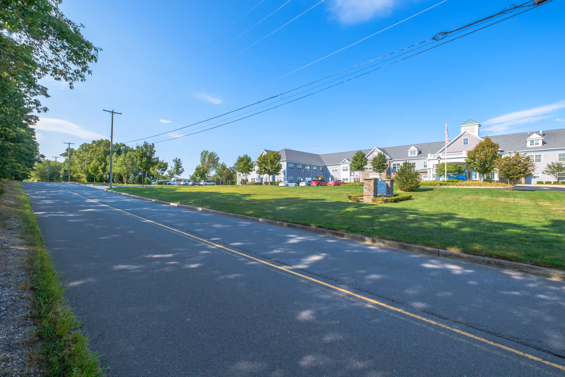 Wide paved road with a double yellow line running alongside a large, two-story senior living facility building with a gray roof and light-colored exterior. The building is surrounded by a well-maintained green lawn with trees and parked cars. The sky is clear and blue.