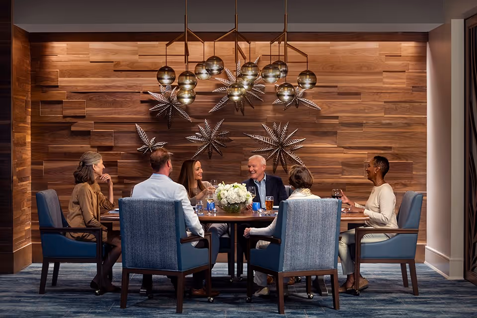 A group of five adults sitting around a round dining table in a warmly lit room with a wooden textured wall decorated with metallic star-shaped ornaments. They are engaged in conversation and appear to be enjoying drinks together. The chairs are upholstered in blue fabric, and a modern chandelier with spherical lights hangs above the table.
