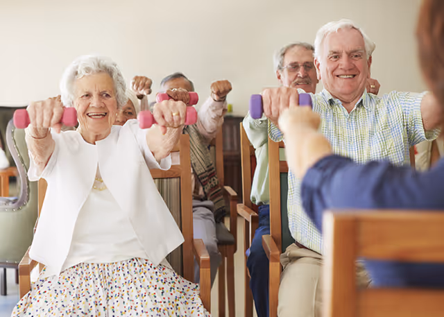 A group of elderly people sitting on chairs in a room, participating in a seated exercise class using small hand weights, led by an instructor.