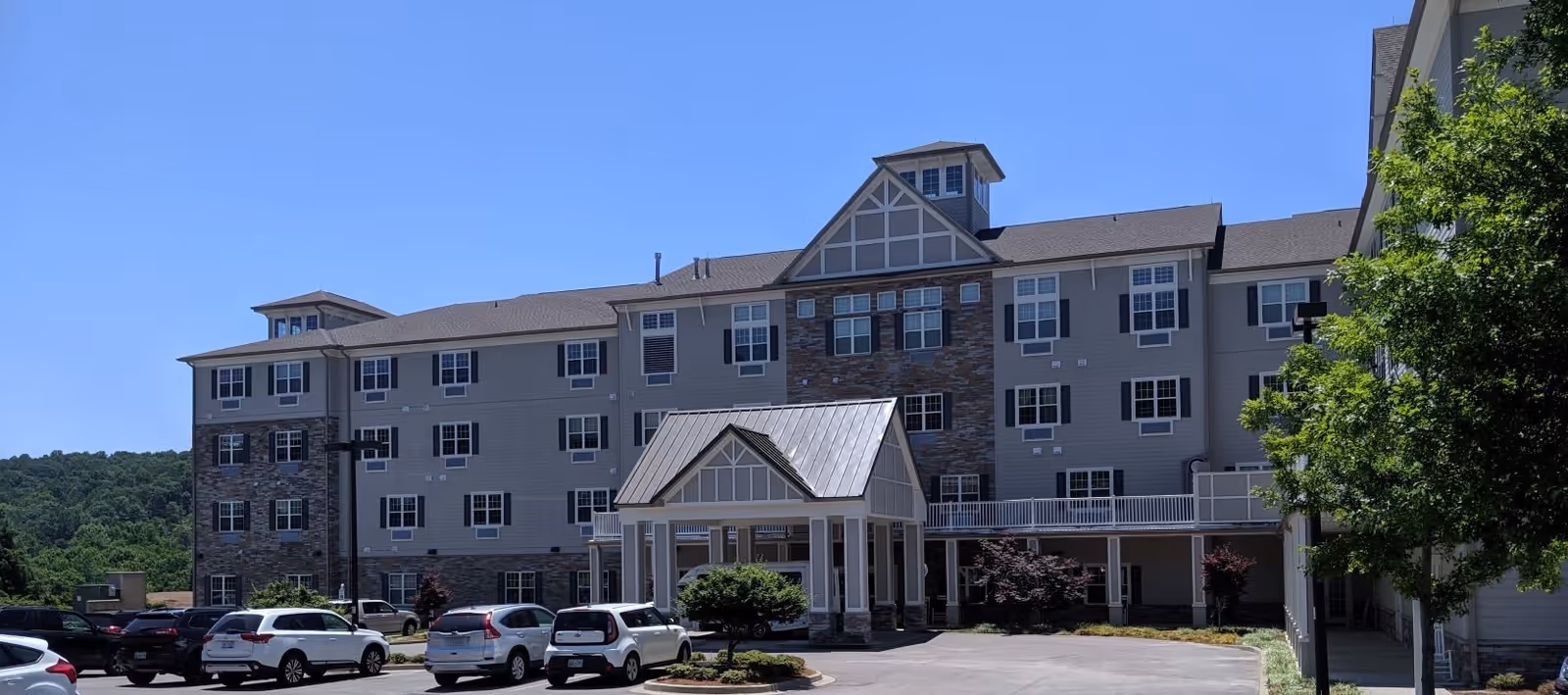 Exterior view of a multi-story senior living facility building with a covered entrance, several parked cars in front, and trees on the right side under a clear blue sky.