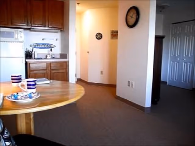 Interior view of a small kitchen area with wooden cabinets, a white refrigerator, microwave, and a sink. In the foreground, there is a round wooden table with two striped mugs and a plate with snacks. The background shows a hallway with a clock on the wall and a partially open door leading to another room.