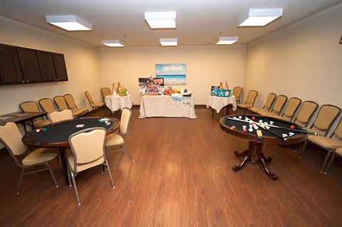 A spacious room with wooden flooring and beige walls, featuring two round tables with chairs around them. One table has poker chips and cards, while the other has billiard balls and cues arranged on it. Along the walls, there are multiple beige chairs lined up. At the far end of the room, there are two small tables covered with white cloths displaying decorative items and a painting of a beach scene on the wall above them.