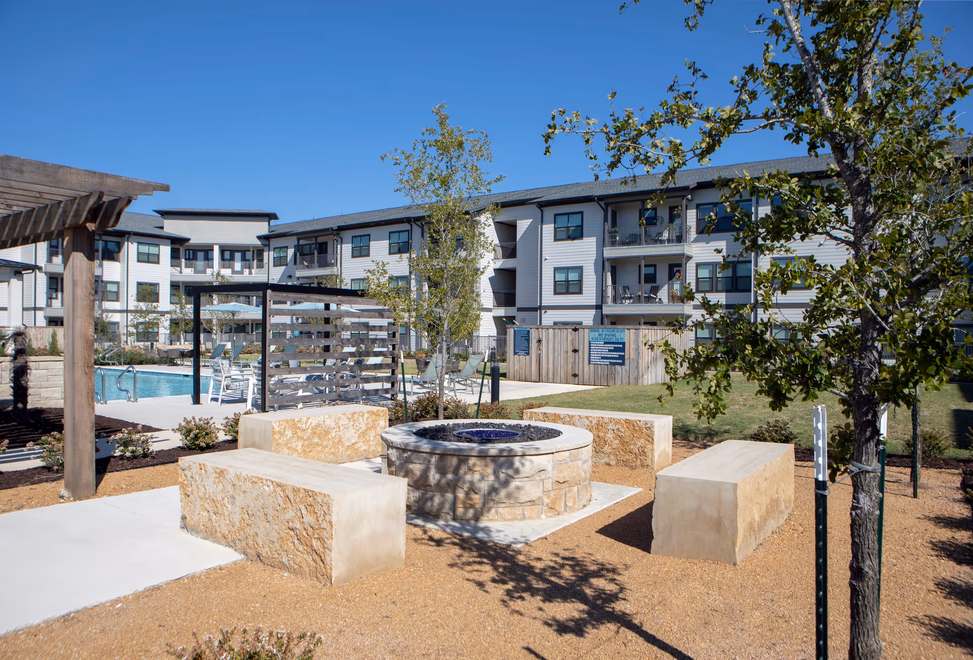 Outdoor seating area with stone benches arranged around a circular fire pit, surrounded by trees and landscaping. In the background, there is a swimming pool with lounge chairs and a three-story residential building under a clear blue sky.