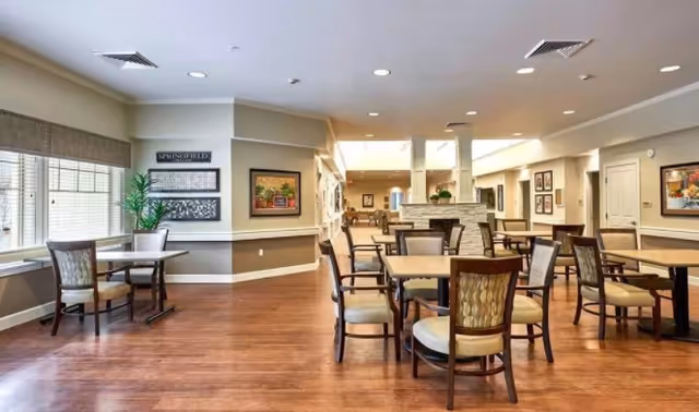 A spacious and well-lit dining area with multiple wooden tables and cushioned chairs arranged neatly on a wooden floor. Large windows on the left side allow natural light to fill the room. The walls are decorated with framed artwork and a sign that reads 'SPRINGFIELD'. The ceiling has recessed lighting and air vents.