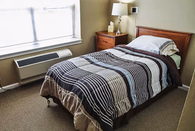 Small bedroom with a single bed dressed in striped bedding, a wooden headboard and nightstand with lamp beside a window and wall-mounted air conditioner.