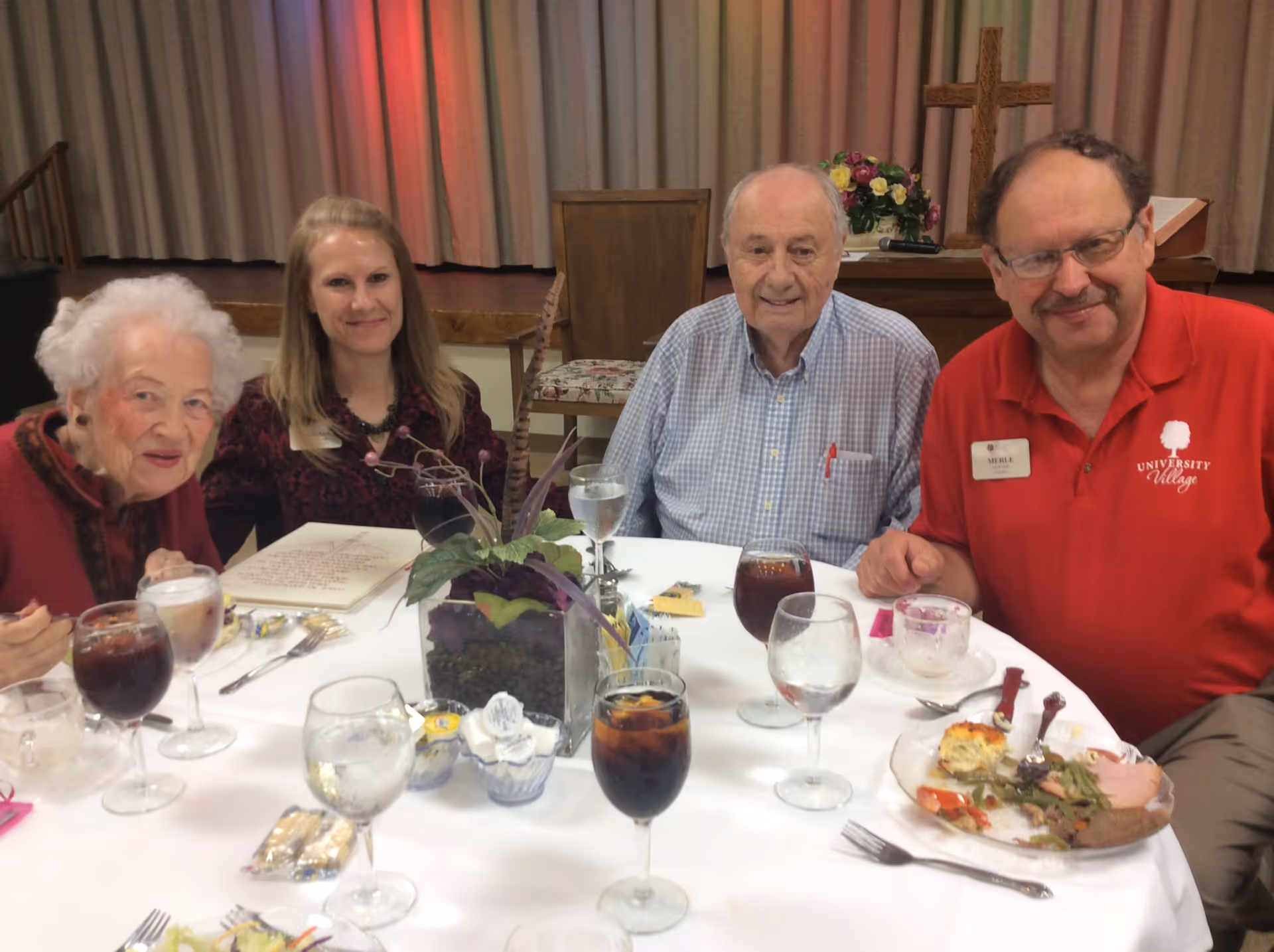 Four people sit around a table with plates, glasses, and a centerpiece at a communal dining event inside a retirement community.