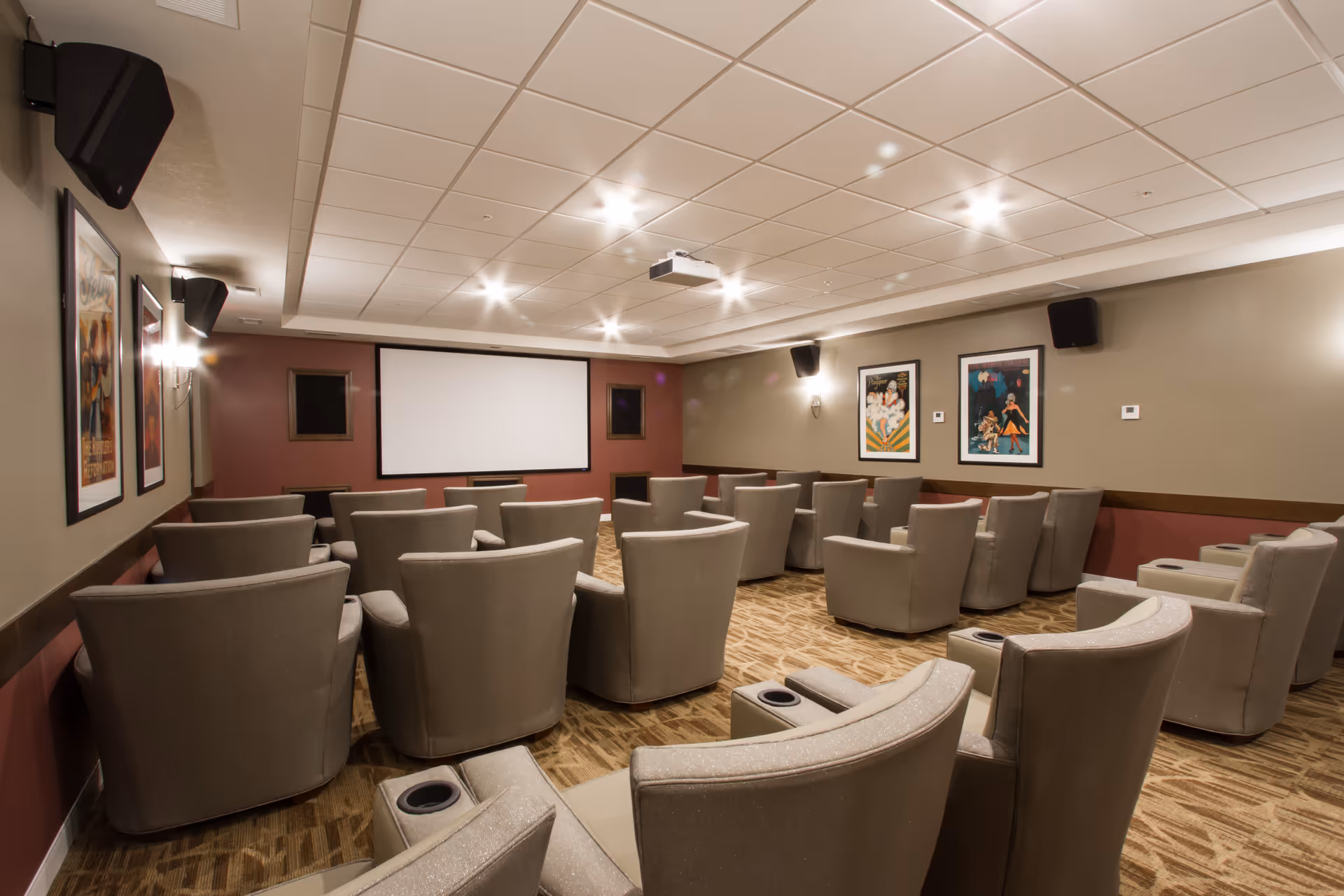 A cozy theater room with multiple rows of comfortable beige armchairs facing a large white projection screen. The room has a carpeted floor, beige and maroon walls adorned with framed artwork, ceiling-mounted speakers, and recessed lighting.