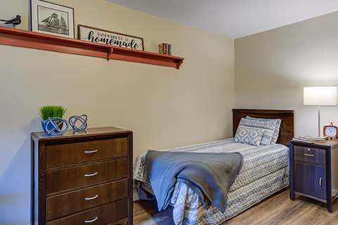 A cozy bedroom with a single bed covered in a patterned bedspread and a gray throw blanket. Next to the bed is a wooden nightstand with a lamp and a red alarm clock. On the opposite side, there is a wooden dresser with three drawers, decorated with two small blue geometric sculptures and a small green plant. Above the dresser, a red wall shelf holds framed pictures, books, and a sign that reads 'A multitude is homemade.' The room has light-colored walls and wood flooring.