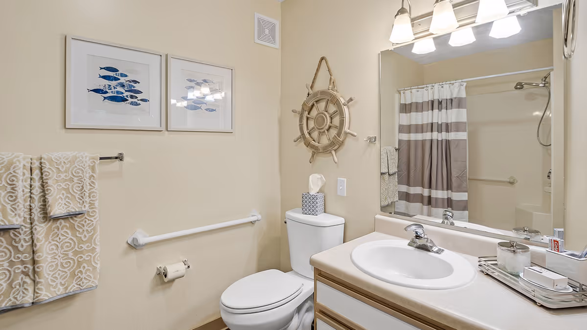 A clean bathroom with beige walls featuring a white toilet, a sink with a countertop, and a large mirror above the sink. There are two framed pictures of blue fish on the wall, a decorative ship wheel hanging next to the mirror, a towel rack with patterned towels, and a shower with a striped curtain in the background.