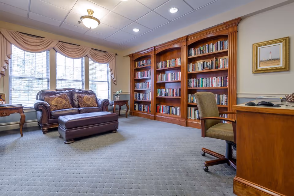 A cozy living room area in Bellamy Fields Assisted Living featuring a large wooden bookshelf filled with books, a brown leather loveseat with two decorative pillows, a matching leather ottoman, two wooden side tables with a small plant, a wooden desk with a computer mouse and keyboard, and an office chair. The room has large windows with beige curtains and a framed picture on the wall.