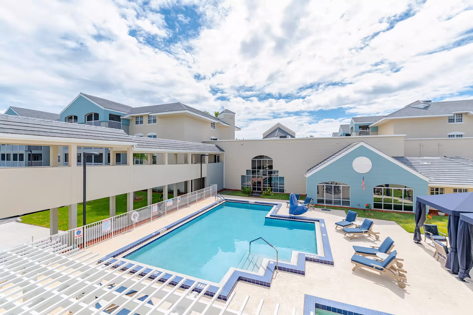 Outdoor swimming pool area at Discovery Village At Boynton Beach with lounge chairs, a shaded cabana, and surrounding buildings under a partly cloudy sky.