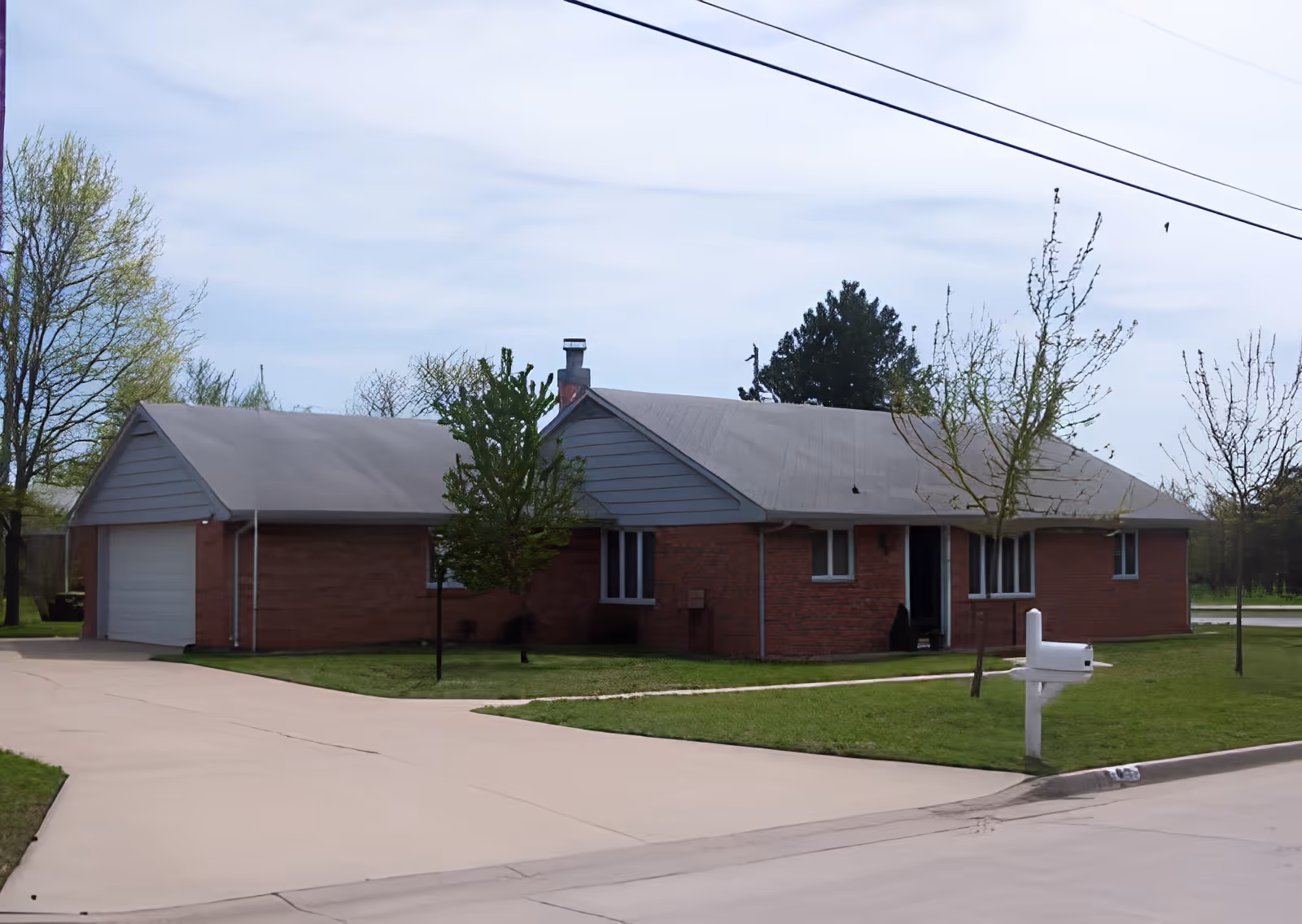 Single-story brick building with a gray roof and attached garage, surrounded by a green lawn with a few small trees and a white mailbox near the street under a partly cloudy sky.