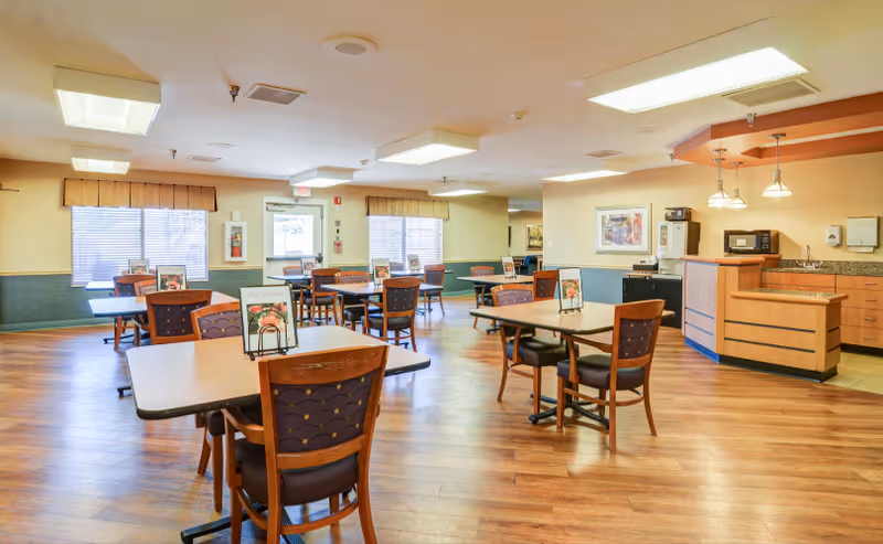A bright dining room with multiple tables and chairs arranged neatly. Each table has a small menu or flyer on it. The room features large windows with blinds and valances, wood flooring, and a small kitchenette area with a microwave, sink, and cabinets. Ceiling lights illuminate the space.