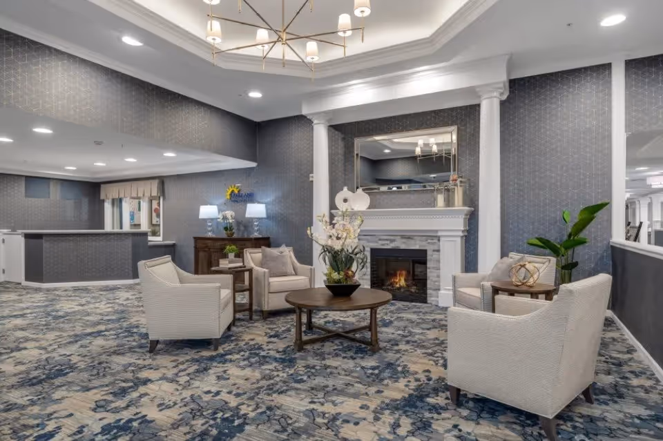 A cozy lounge area with four beige armchairs arranged around a round wooden coffee table with a flower arrangement. Behind the seating area is a white fireplace with a lit fire and a large mirror above it. The walls are decorated with a subtle geometric pattern wallpaper, and the floor is covered with a blue and gray patterned carpet. There is a wooden sideboard with two lamps and a plant, and a reception desk is visible in the background.