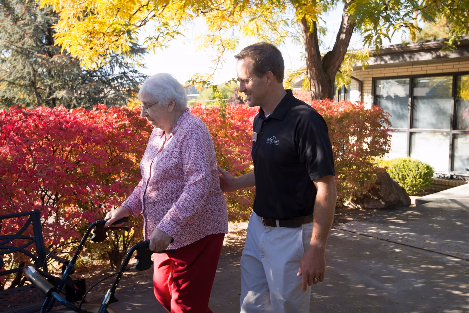 An elderly woman using a walker is being assisted by a man wearing a black polo shirt with a Highland Care Center logo. They are outdoors on a sunny day, surrounded by vibrant red and yellow autumn foliage near a building.