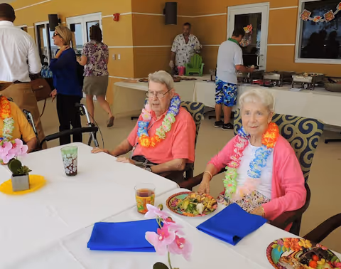 Two elderly women wearing colorful leis sitting at a table with plates of food and blue napkins in front of them. In the background, several people are standing near a buffet table with food trays. The setting appears to be an indoor or covered patio area with yellow walls and windows.
