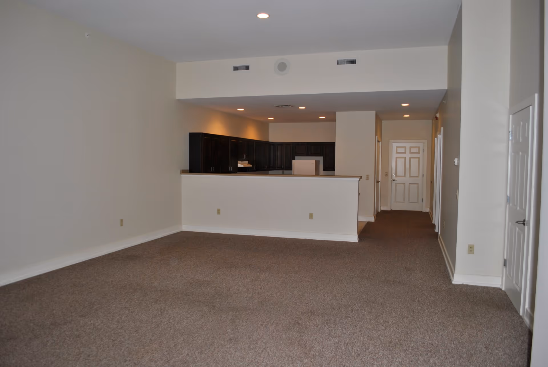 An empty apartment interior showing a large carpeted living area with beige walls and ceiling lights. In the background, there is a kitchen with dark wood cabinets, a white refrigerator, and a half wall separating the kitchen from the living space. A hallway with multiple doors leads to other rooms.