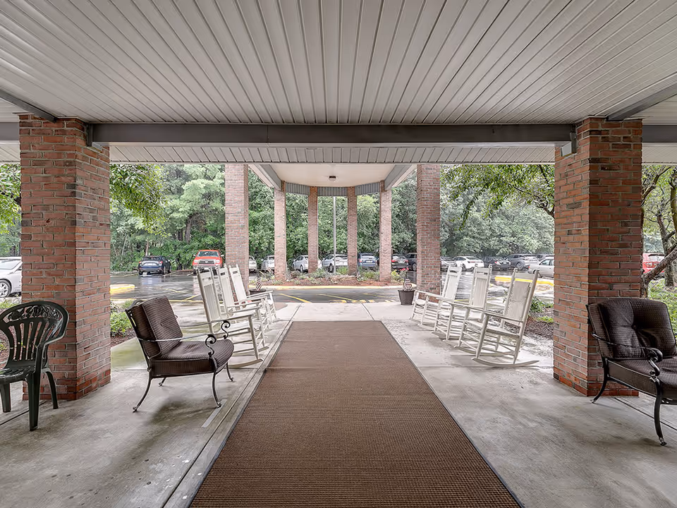 Covered outdoor seating area with several white rocking chairs and two brown cushioned chairs arranged along brick pillars, overlooking a parking lot with cars and surrounded by trees.