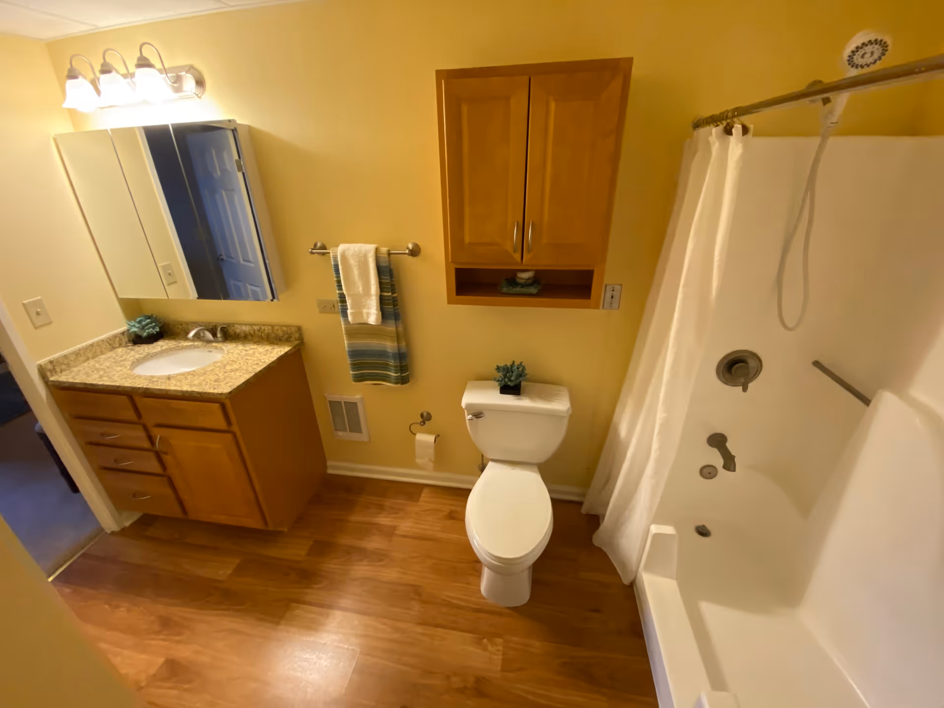 A bathroom with wooden floor and yellow walls featuring a white toilet with a small plant on top, a bathtub with a white shower curtain, a wooden cabinet mounted on the wall above the toilet, a granite countertop with a sink and wooden vanity with drawers, a mirror with lights above it, and a towel rack with two towels.