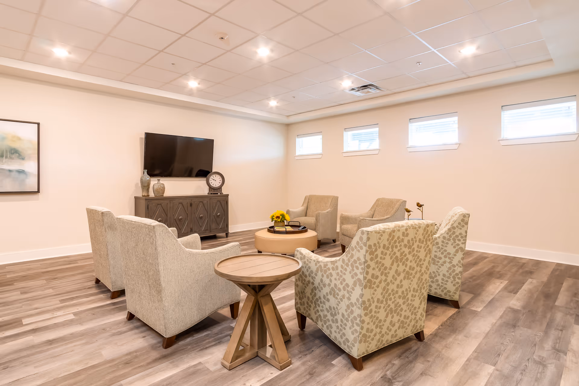 Bright lounge with upholstered armchairs arranged around a coffee table facing a wall-mounted TV.