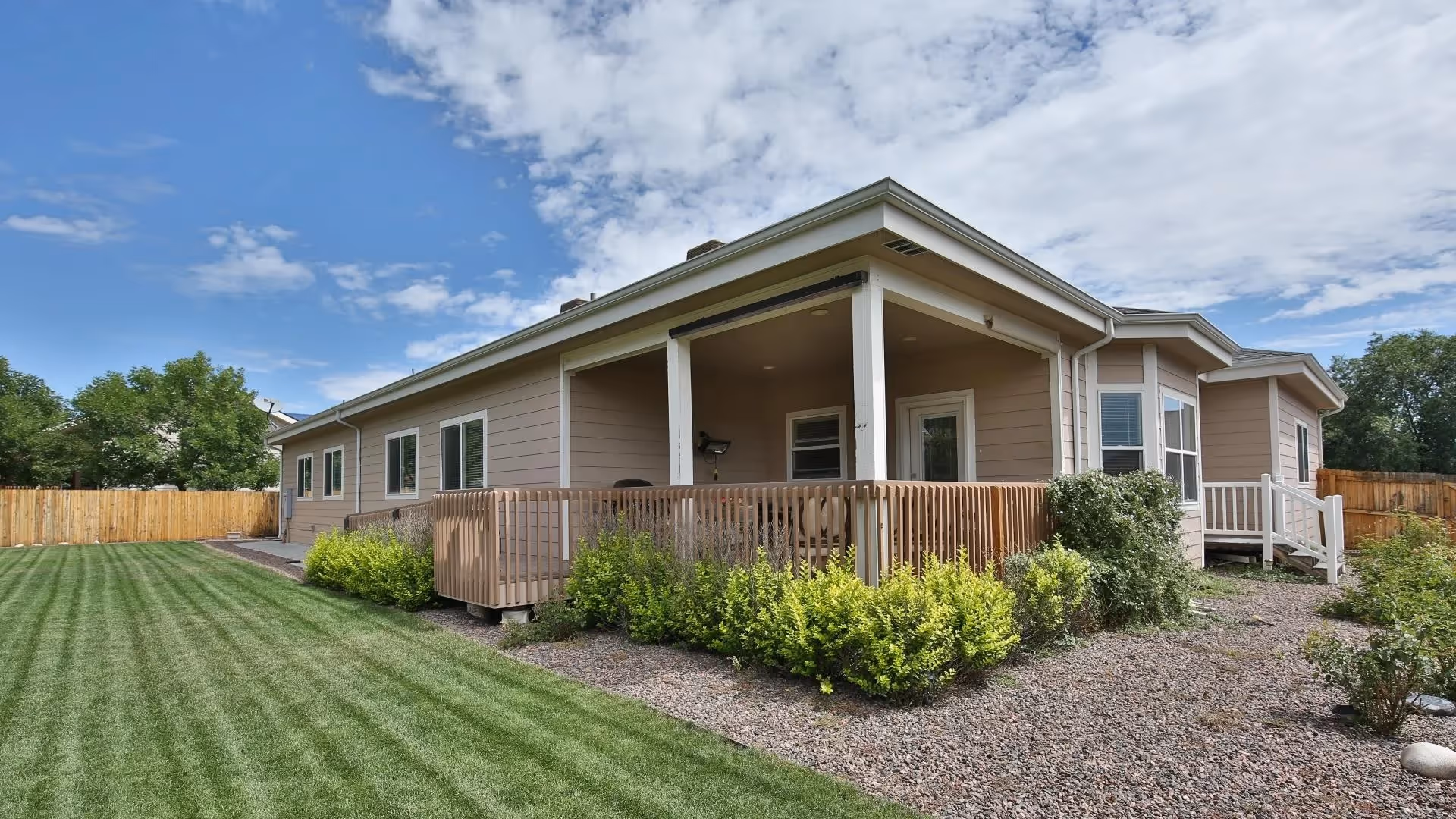 Exterior view of a single-story assisted living facility building with beige siding, a covered porch with wooden railing, surrounded by a well-maintained lawn, shrubs, and a wooden fence under a partly cloudy sky.