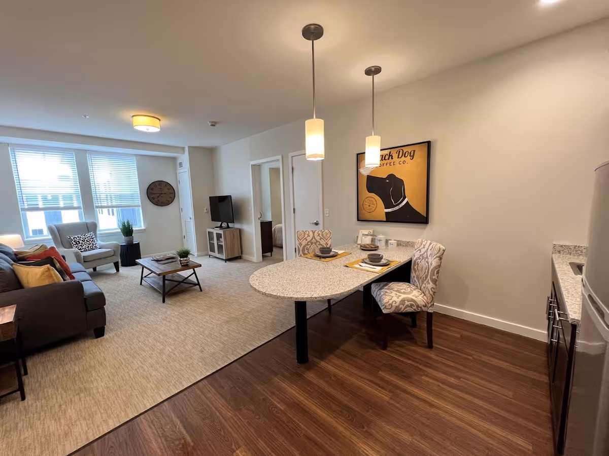 A cozy living and dining area in a senior living facility apartment. The living room features a gray sofa with colorful pillows, a light gray armchair, a wooden coffee table, a TV on a stand, and a large wall clock. The dining area has a small oval table set for two with patterned chairs and two pendant lights hanging above. The floor transitions from carpet in the living area to wood in the dining and kitchen area. A framed picture of a black dog with the text 'Black Dog Coffee Co.' hangs on the wall.