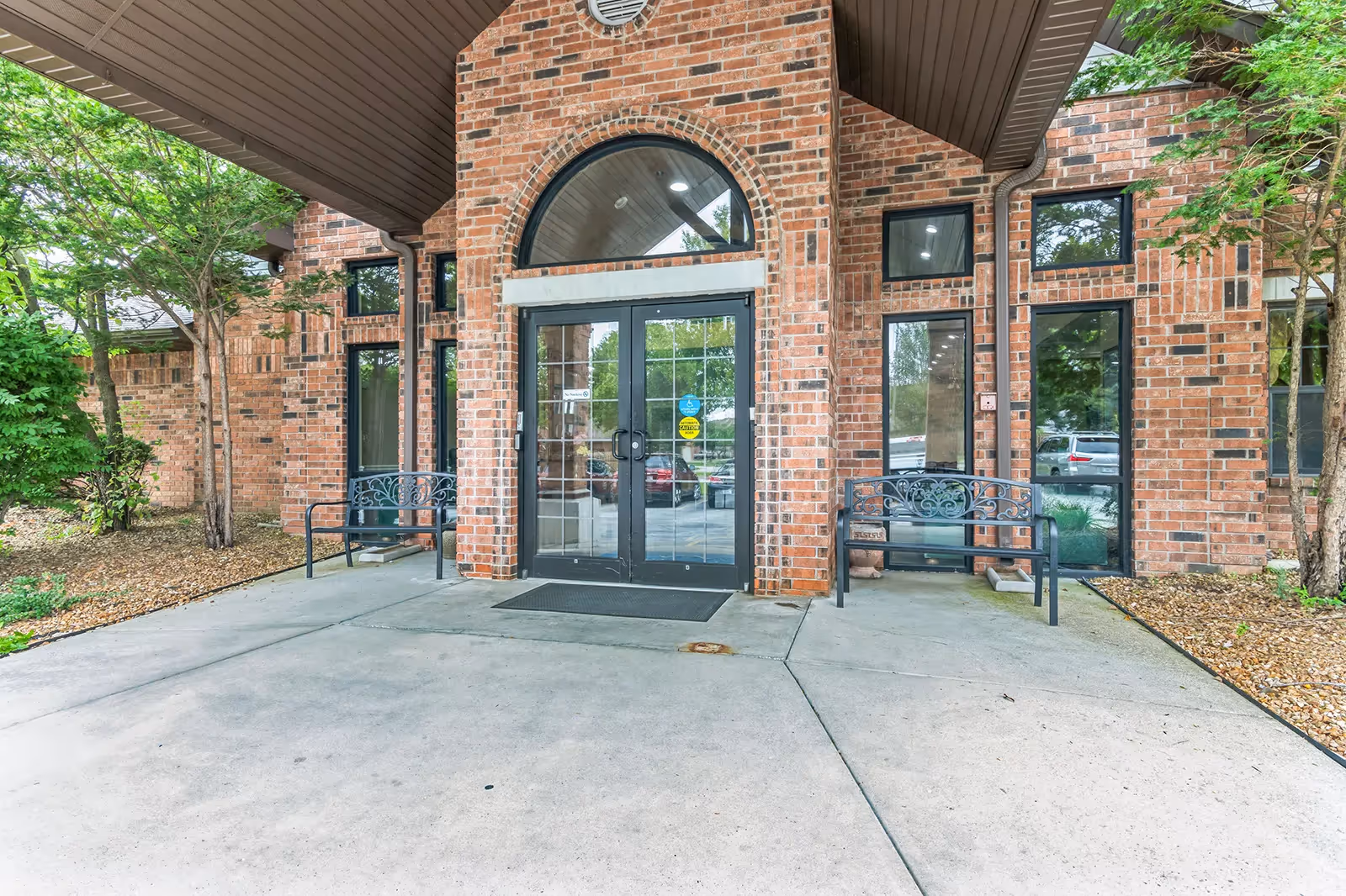 Entrance to a brick building with double glass doors under a covered area. Two black metal benches are placed on either side of the entrance. There are tall windows on both sides of the doors and small trees and landscaping around the building.