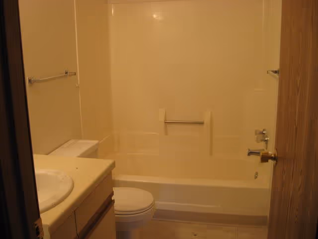 A small bathroom with a white bathtub and shower combination, a toilet, and a sink with a countertop. The walls and bathtub surround are light-colored, and there are towel bars on both sides of the bathtub. The door to the bathroom is partially open.