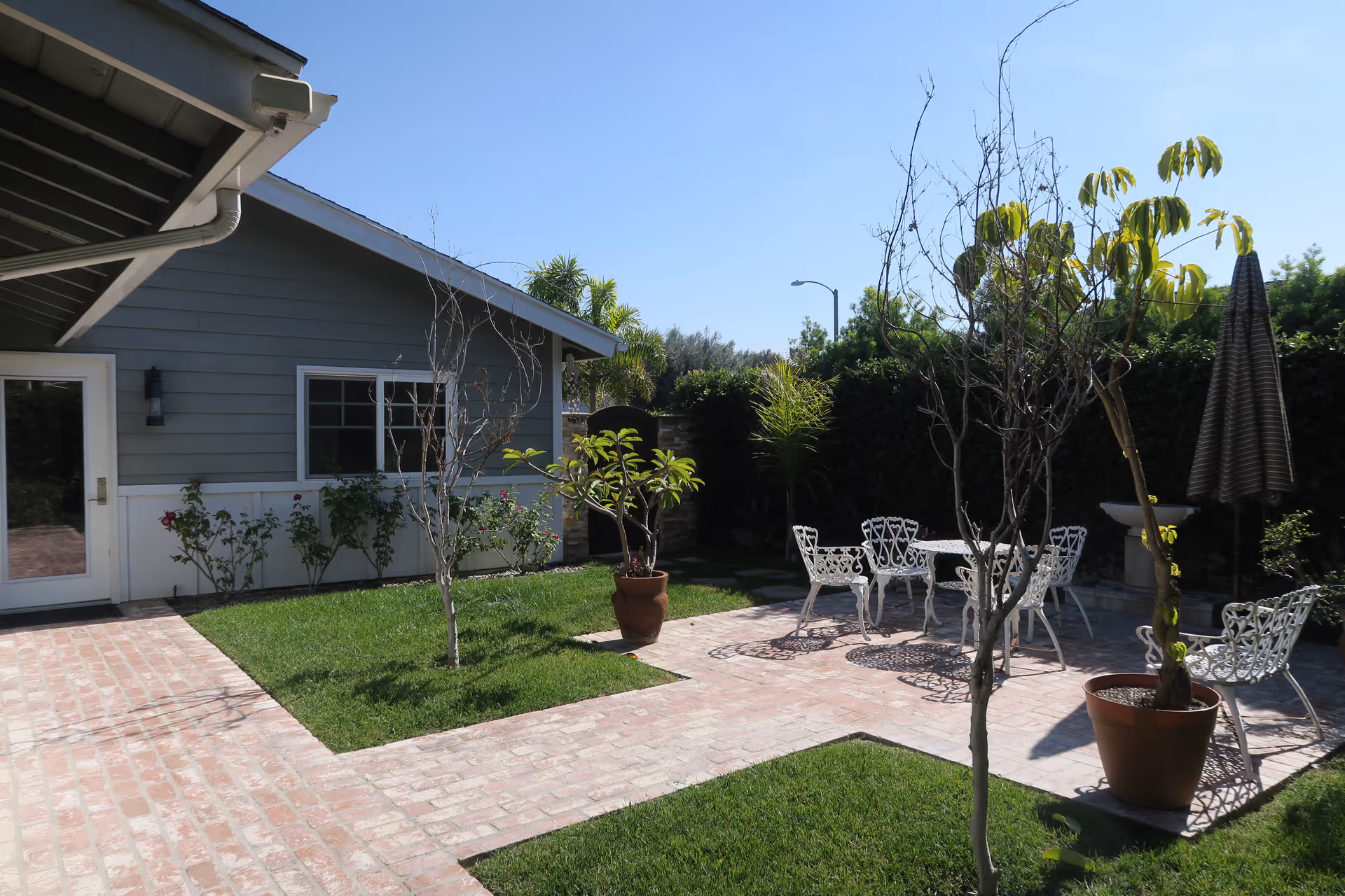 Sunlit backyard patio with brick walkways, potted plants and white metal table and chairs next to a gray single-story house.