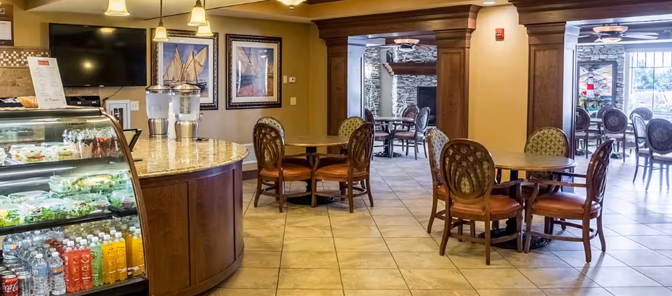 Interior view of a senior living facility dining area with round wooden tables and cushioned chairs. On the left side, there is a curved counter with a glass display case containing bottled drinks and packaged food. The walls are decorated with framed pictures, and there is a TV mounted above the counter. The room has tiled flooring and warm lighting, with an adjacent seating area visible through a wide doorway.