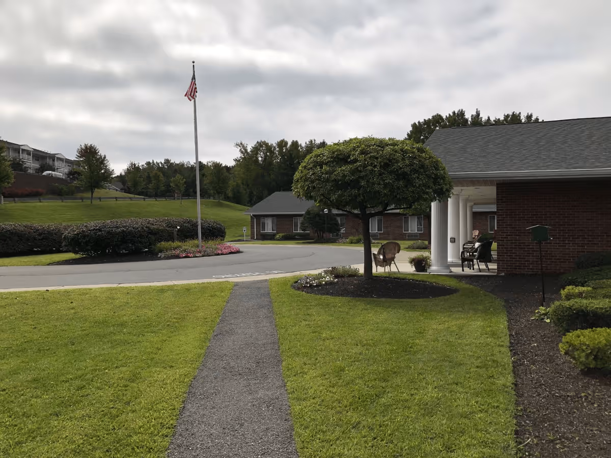 Outdoor view of a senior living facility with a paved walkway leading to a circular driveway. There is a flagpole with an American flag, well-maintained green lawns, bushes, and a tree with a rounded canopy. A brick building with white columns and outdoor seating is visible on the right side, with a person sitting on a bench waving.