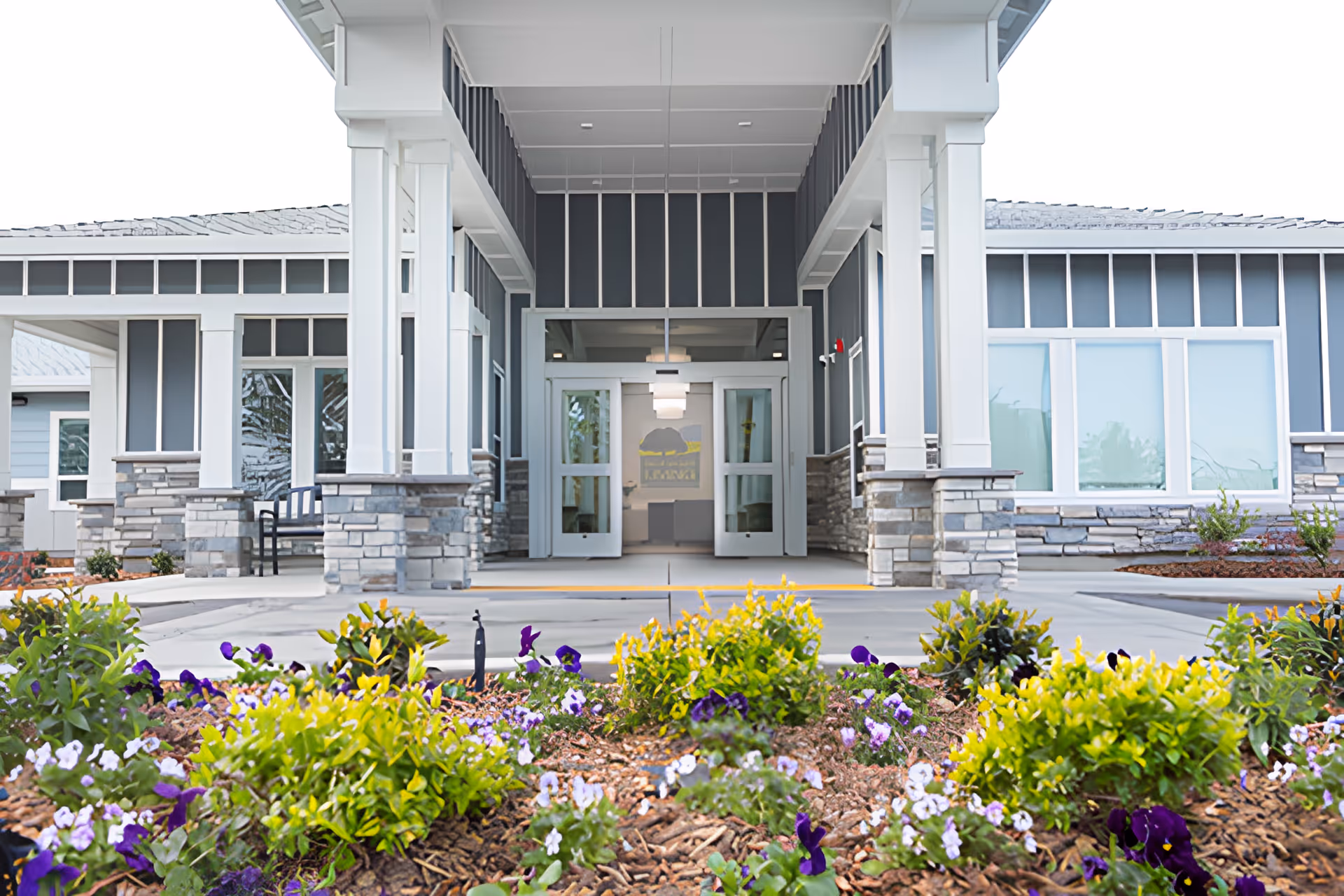Front entrance of a single-story memory-care building with a covered entryway, stone-clad columns, glass doors, and landscaped flower beds in the foreground.