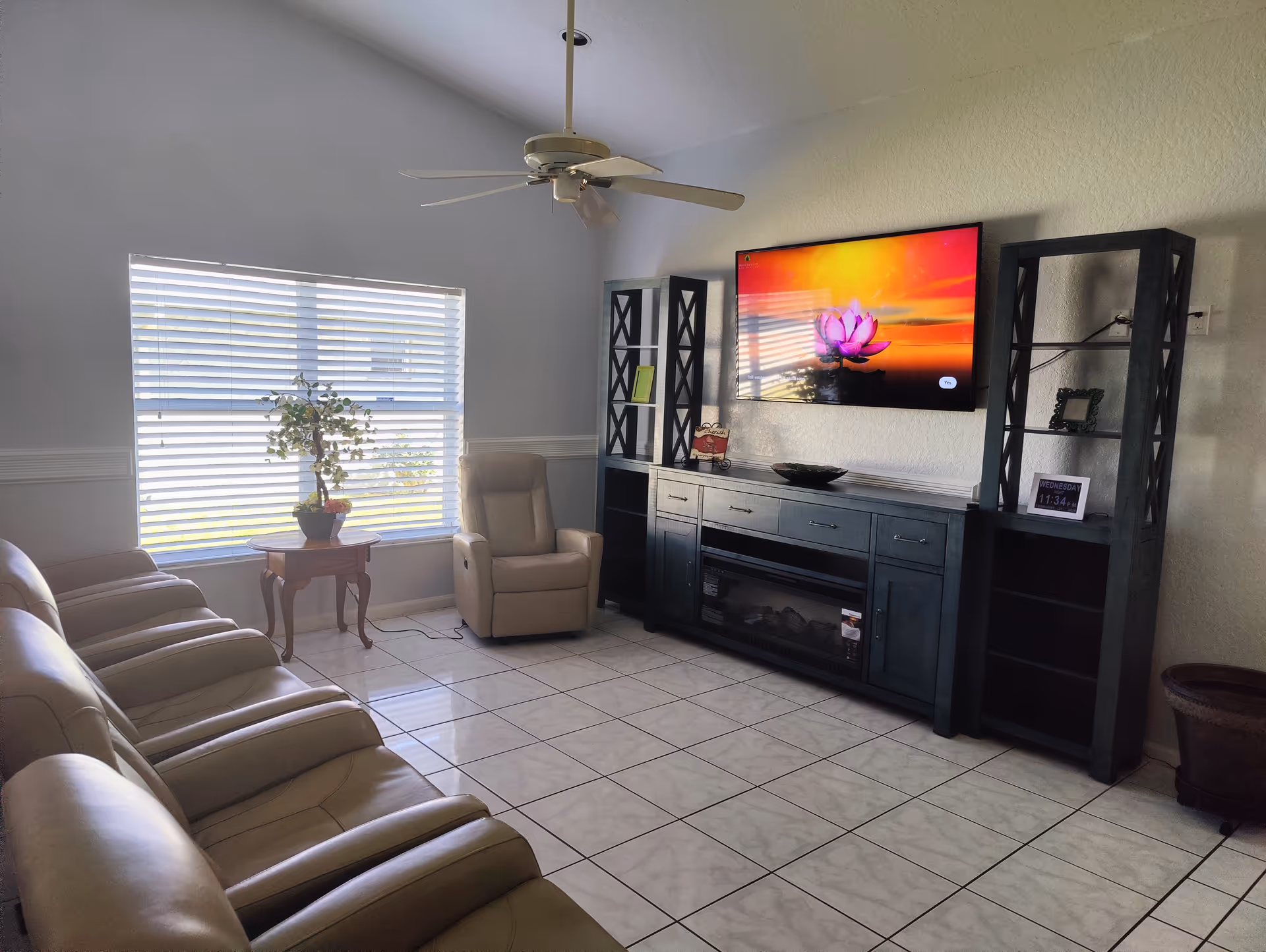 A bright living room with a row of beige leather recliners facing a wall-mounted flat screen TV displaying a colorful lotus flower image. The room has white tiled floors, a ceiling fan, a large window with blinds, a small wooden side table with a potted plant, and black shelving units flanking a black entertainment console with a built-in electric fireplace.