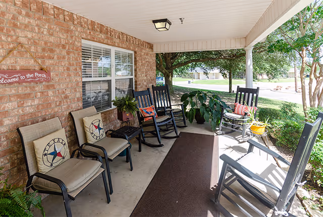 Covered porch area with several cushioned chairs and rocking chairs arranged around a brown outdoor rug. The porch has a brick wall with a window and a sign that reads 'Welcome to the Porch'. There are plants and trees visible around the porch, providing a shaded and inviting outdoor seating space.