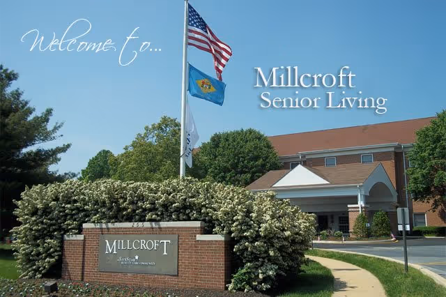 Entrance to Millcroft Senior Living facility with a brick sign surrounded by flowering bushes, three flags on a flagpole including the American flag, and a large building with a covered entrance in the background under a clear blue sky.