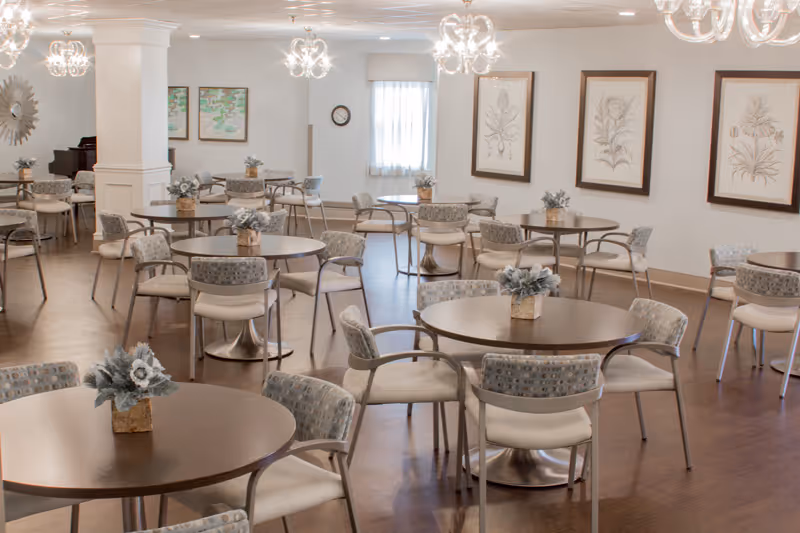 Bright dining/activity room with round tables and chairs, floral centerpieces, chandeliers and framed botanical art on the walls.