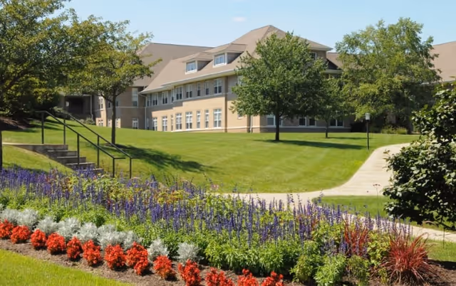 Exterior view of a senior living facility building with a manicured lawn, trees, and colorful flower beds.