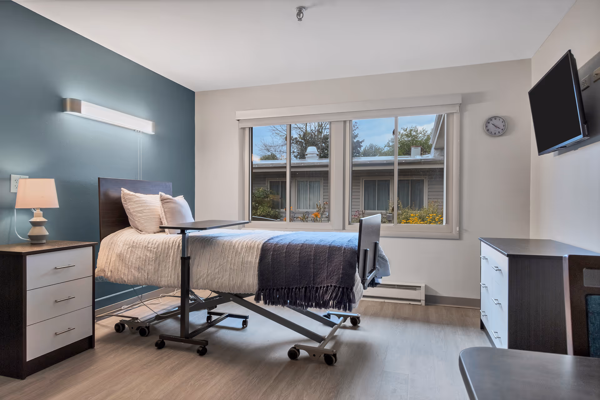 A bright and clean bedroom in a senior living facility with a hospital-style adjustable bed covered with white and dark blue bedding. The room has a large window showing an exterior view of another building and some greenery. There is a bedside table with a lamp, a wall-mounted light above the bed, a clock on the wall, a flat-screen TV mounted on the wall, and a dresser with drawers.