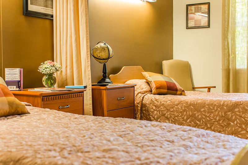 A cozy bedroom with two twin beds covered in beige patterned bedspreads. Each bed has a colorful pillow. Between the beds is a wooden nightstand with a globe on it. There is a vase with flowers and some books on another wooden piece of furniture. The room has beige walls, a framed picture, and curtains letting in natural light.