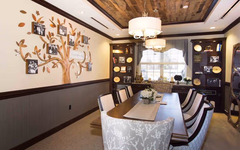 A well-lit dining room with a long wooden table surrounded by upholstered chairs. The walls feature dark wood paneling on the lower half and light paint above, decorated with a large tree-shaped photo display holding black and white pictures. Two chandeliers hang from a wooden ceiling. Shelves with decorative items and framed photos flank a window with blinds and a valance.