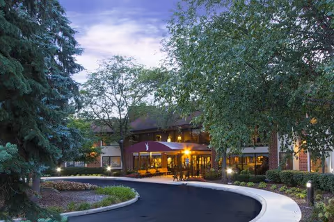 Exterior view of a senior living facility at dusk with a curved driveway leading to the entrance. The building is surrounded by trees and landscaping, with warm lights illuminating the entrance area.