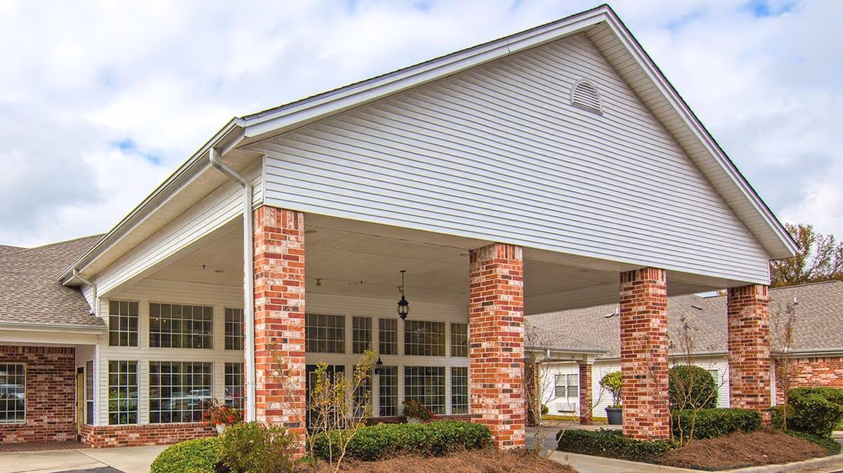 Exterior view of a senior living facility with a large covered entrance supported by brick columns. The building features white siding and multiple windows, surrounded by landscaped bushes and plants under a partly cloudy sky.