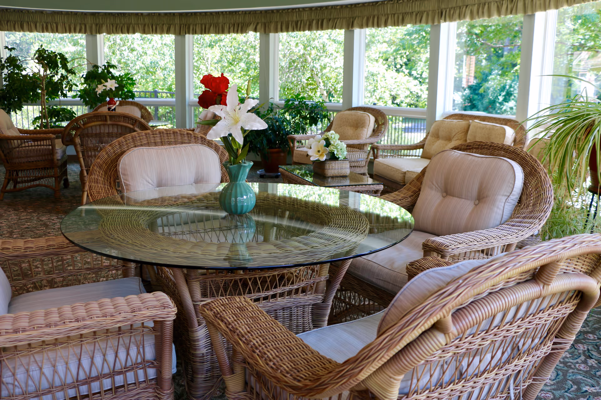 Sunroom with wicker furniture, a glass-top round table, cushioned chairs and potted plants visible through large windows.
