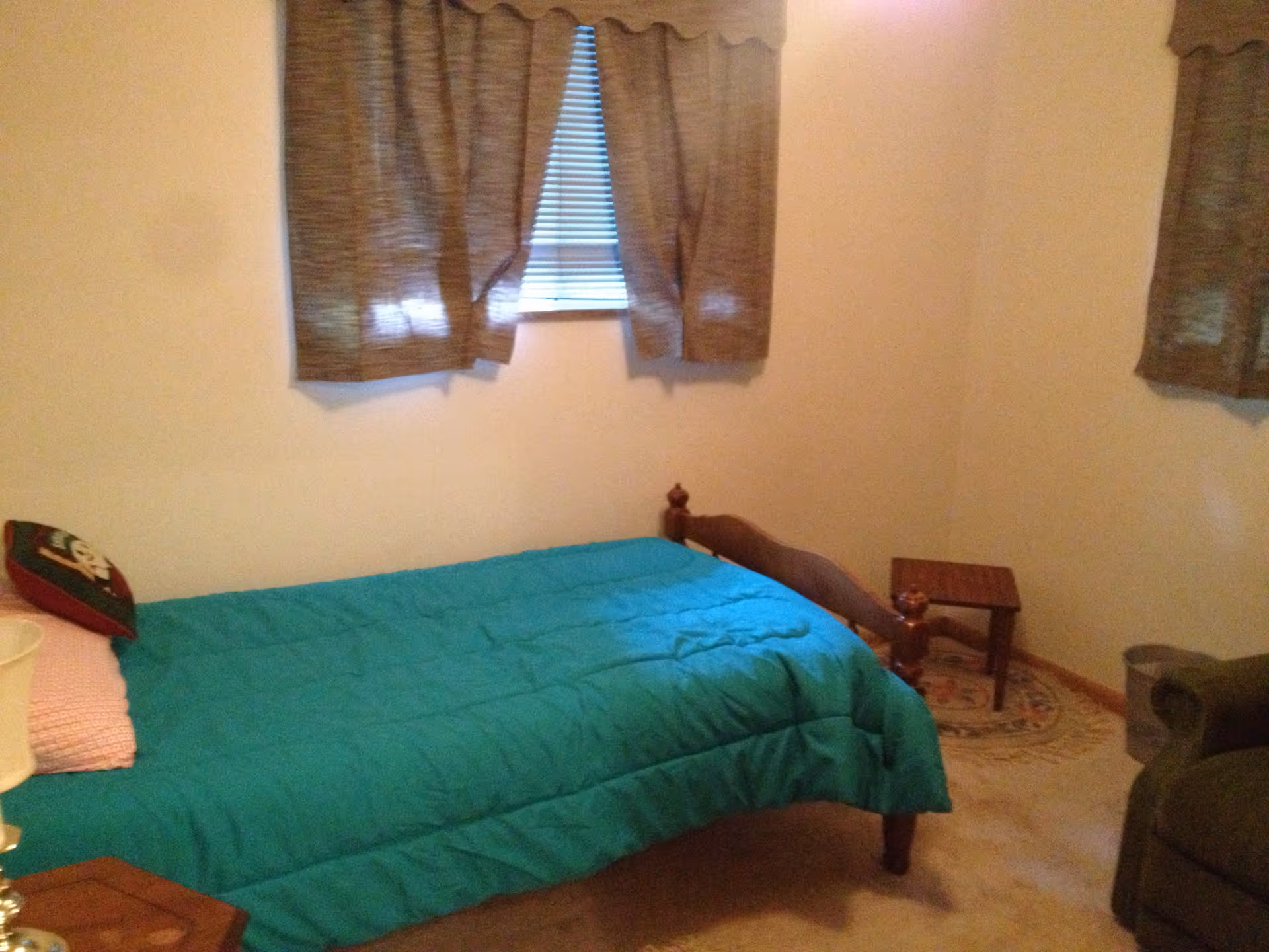Small bedroom with a single wooden bed covered by a teal comforter, brown curtains over a window, and a small side table.